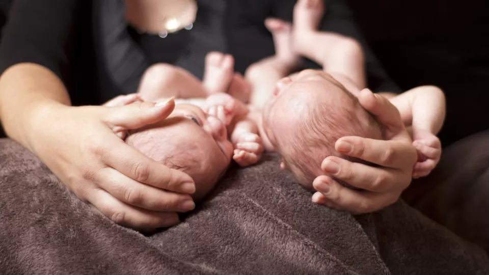 Ett par nakna nyfödda tvillingar ligger på en brun mjuk filt med huvudena vända mot kameran och samtidigt snett mot varandra. Varsamt på varsitt huvud vilar en vuxen hand som med omsorg omfamnar de små sköra liven.