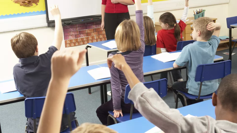 Barn i sina skolbänkar som räcker upp händerna. Foto