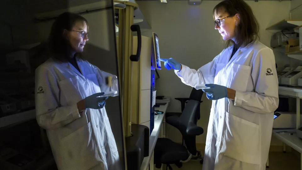 Forskare i vit labbrock tittar in i en maskin i ett mörkt rum. Hennes spegelbild reflekteras i en glasruta. Foto.