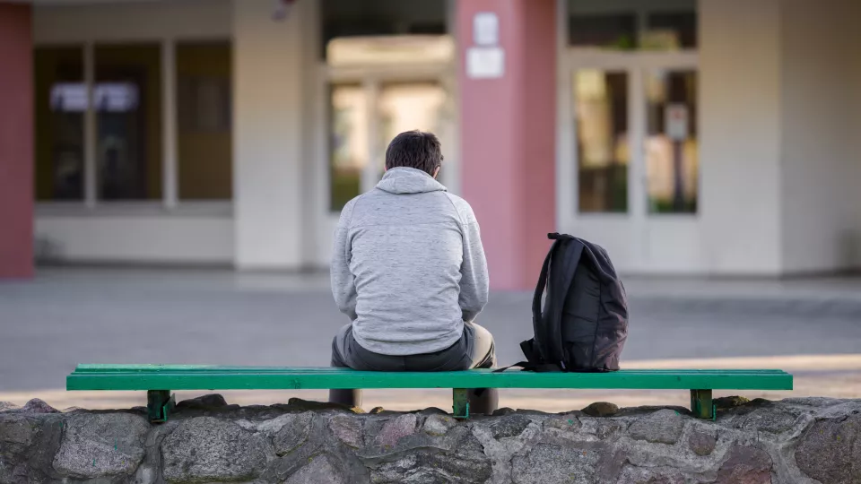 Ensam ung man sitter på en bänk utanför en skola. har ryggen mot kameran. Foto: iStock FotoDuets 