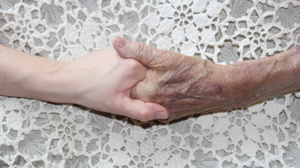 Yngre hand håller i en äldre hand, båda vilar mot ett vitt, virkat överkast.