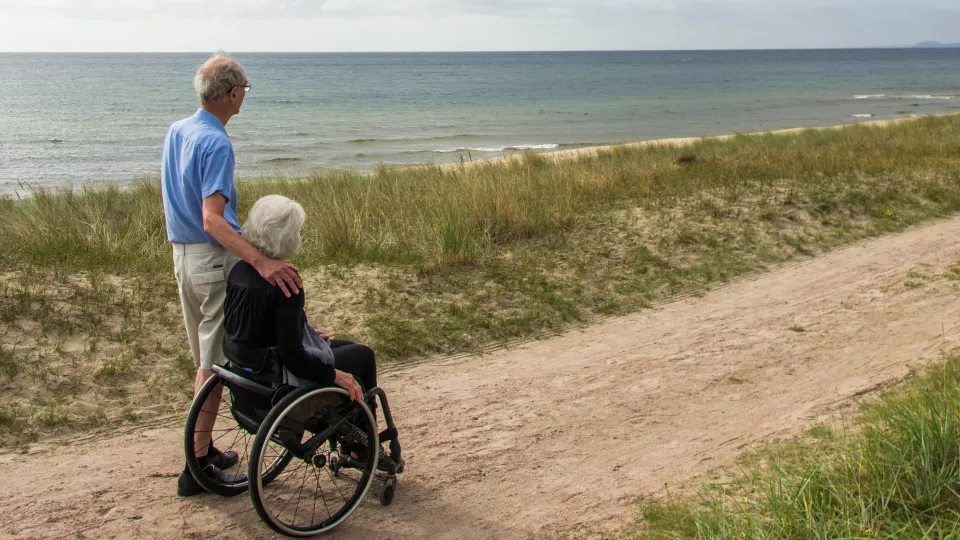 Äldre par på stranden. Hon i rullstol, han intill med handen på hennes axel. Foto: Sophie Jörgensen.