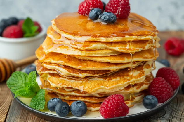 En hög med pannkakor dekorerade med hallon, blåbär och citronmeliss. På kanterna rinner det sirap. Det ser gott ut. Foto.