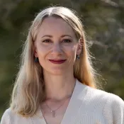 Elin har långt blont hår som glittrar i solen, röda läppar och vit topp.