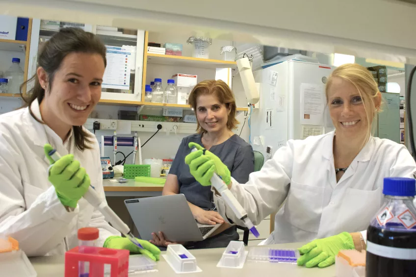 Tre kvinnor i laboratoriemiljö, två har vita rockar och gröna handskar med pipetter i händerna.