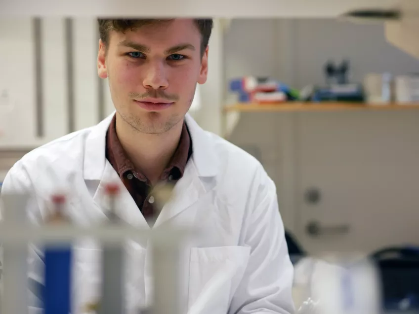 Porträtt på Einar Smith klädd i vit labbrock bakom provrör mellan labbhyllor. 