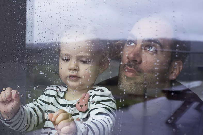 Litet barn och en man tittar ut genom ett regnigt fönster. Foto.